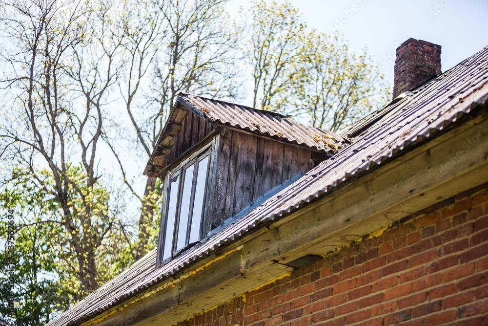 Old wooden roof with small window and chimney in rural Latvia, Europe. Red brick wall weathered with texture. Trees in background. Old wooden roof with small window and chimney in rural Latvia, Europe. Red brick wall weathered with texture. Trees in background.
