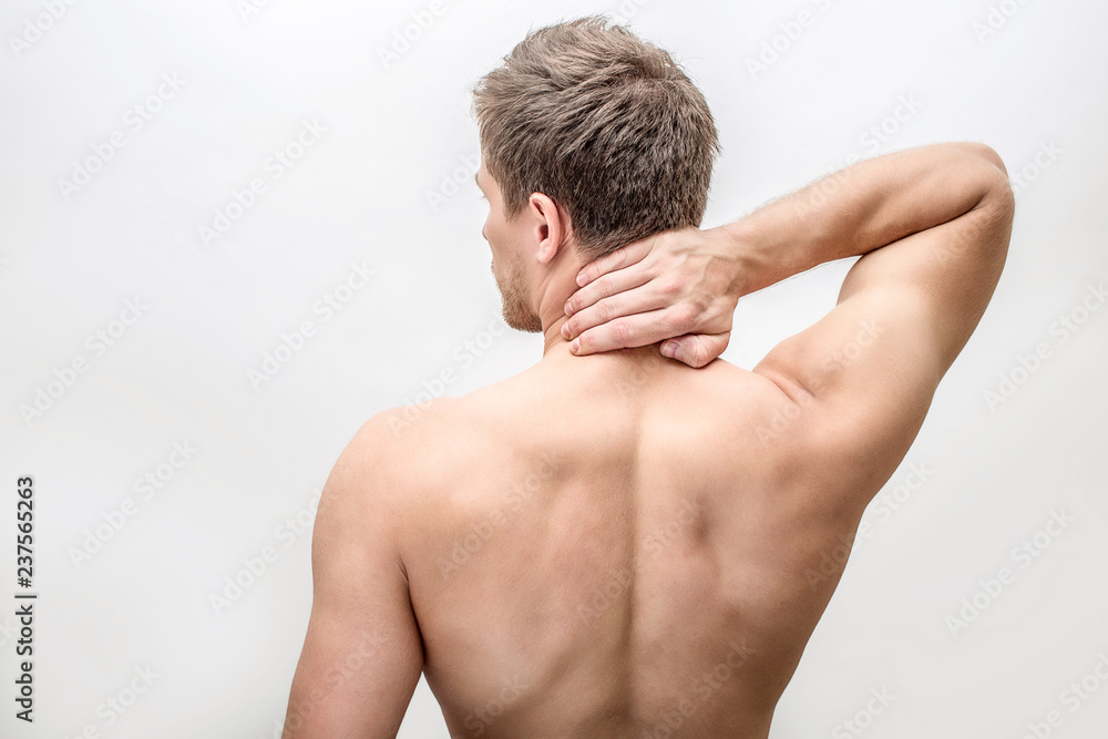 Shirtless young man stand and hold hand on neck. He feels pain there. Guy shows his back. Isolated on white background.