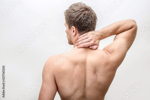 Shirtless young man stand and hold hand on neck. He feels pain there. Guy shows his back. Isolated on white background.