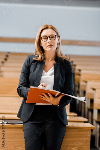 female university professor looking at camera, holding journal and writing in classroom photo