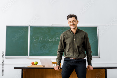 smiling male teacher in formal wear looking at camera and standing near desk in classroom