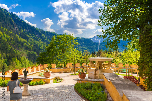 A woman admires the beautiful garden of Hohenschwangau castle with the Lion Fountain, the small Goose Man Fountain, flowers, trees and benches; surrounded by the forest and the Alps in the background. photo
