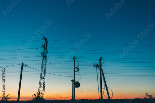 Power lines in field on sunrise background. Silhouettes of poles with wires at dawn. Cables of high voltage on warm orange blue sky. Power industry at sunset. Many cables in picturesque vivid sky.