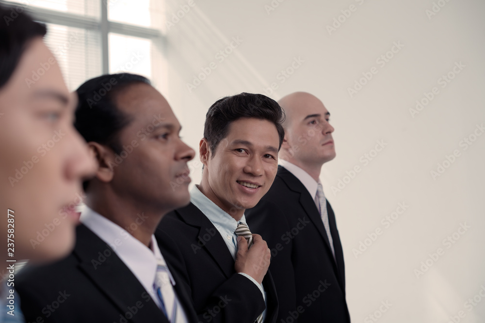 Handsome smiling Vietnamese man adjusting necktie and looking at camera when standing among coworkers at meeting Handsome smiling Vietnamese man adjusting necktie and looking at camera when standing among coworkers at meeting