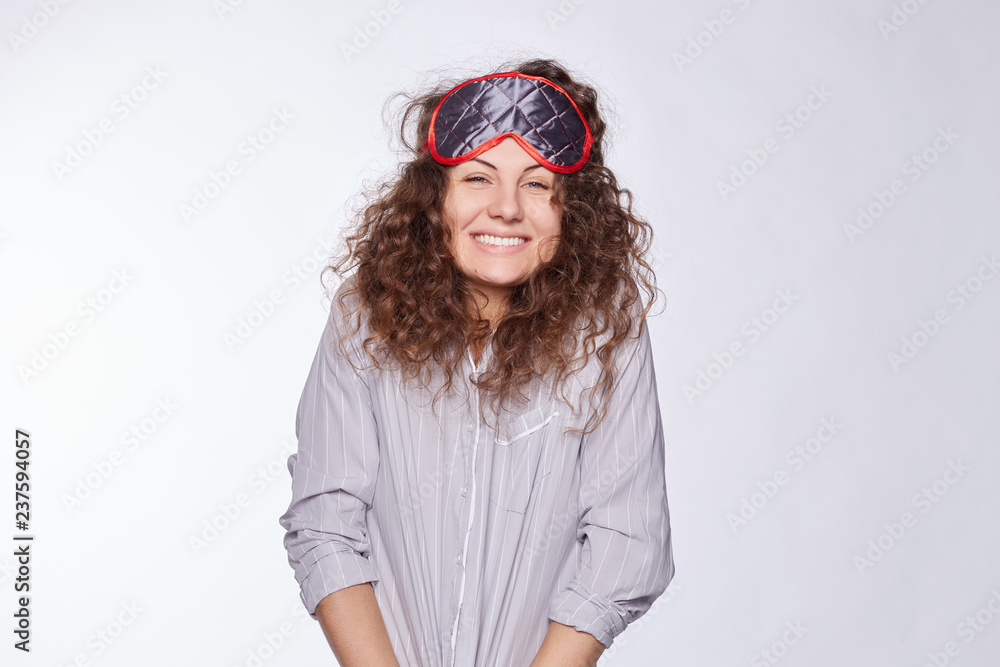 Pleased female glad to receive coffee at bed from lover, dressed in fashionable grey pajamas, has sleeping mask on head, has good sleep, isolated over white wall. Positive emotions early in morning.