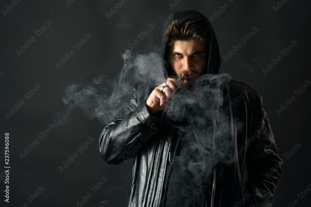 Man with vaping mod exhaling steam at black studio background. Bearded guy smoking e-cigarette to quit tobacco. Vapor and alternative nicotine free smoking concept Man with vaping mod exhaling steam at black studio background. Bearded guy smoking e-cigarette to quit tobacco. Vapor and alternative nicotine free smoking concept