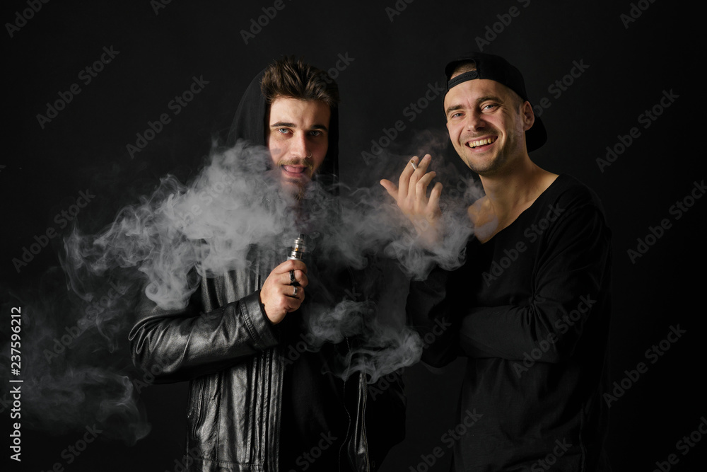 Bad habit, smoking in a public place. Two Young man blowing smoke to join it in one cloud at black studio background. Friends and vape addiction concept. Bad habit, smoking in a public place. Two Young man blowing smoke to join it in one cloud at black studio background. Friends and vape addiction concept.