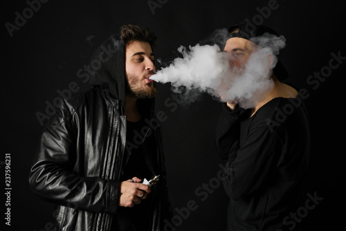 Bad habit, smoking in a public place. Two Young man blowing smoke to join it in one cloud at black studio background. Friends and vape addiction concept. 