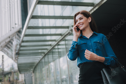 Concept of success and well-being. Waist up low angle portrait of smiling successful lady in stylish cloth talking by smartphone near business center. Copy space on left