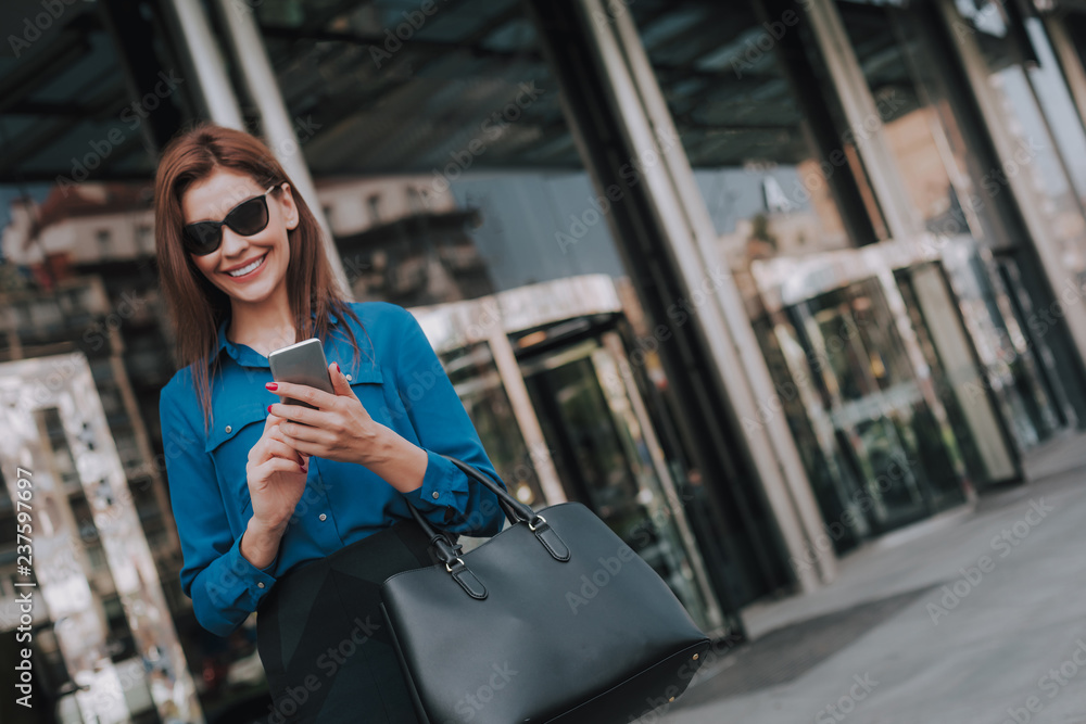 Fototapeta premium Concept of success and well-being. Waist up portrait of smiling business lady in stylish cloth walking on street with mobile phone. Copy space on right
