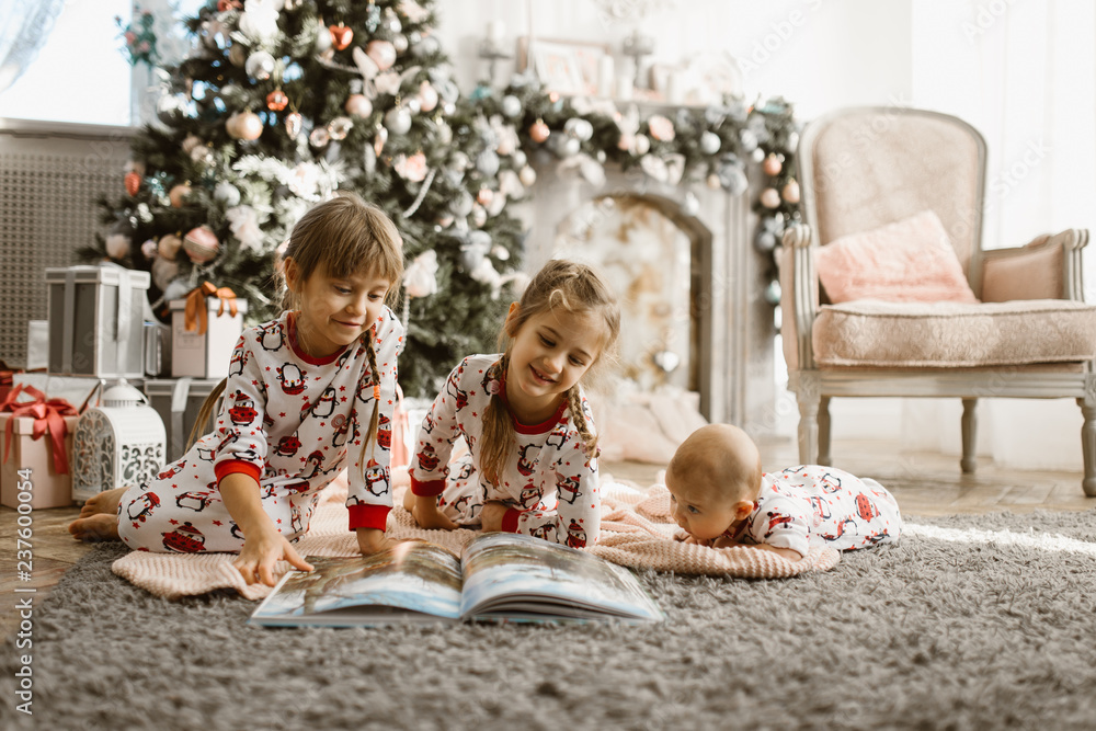 Two little sisters and a tiny brother lie on the carpet and read book near the New Year's tree with gifts in the light cozy room with armchair and fireplace Two little sisters and a tiny brother lie on the carpet and read book near the New Year's tree with gifts in the light cozy room with armchair and fireplace