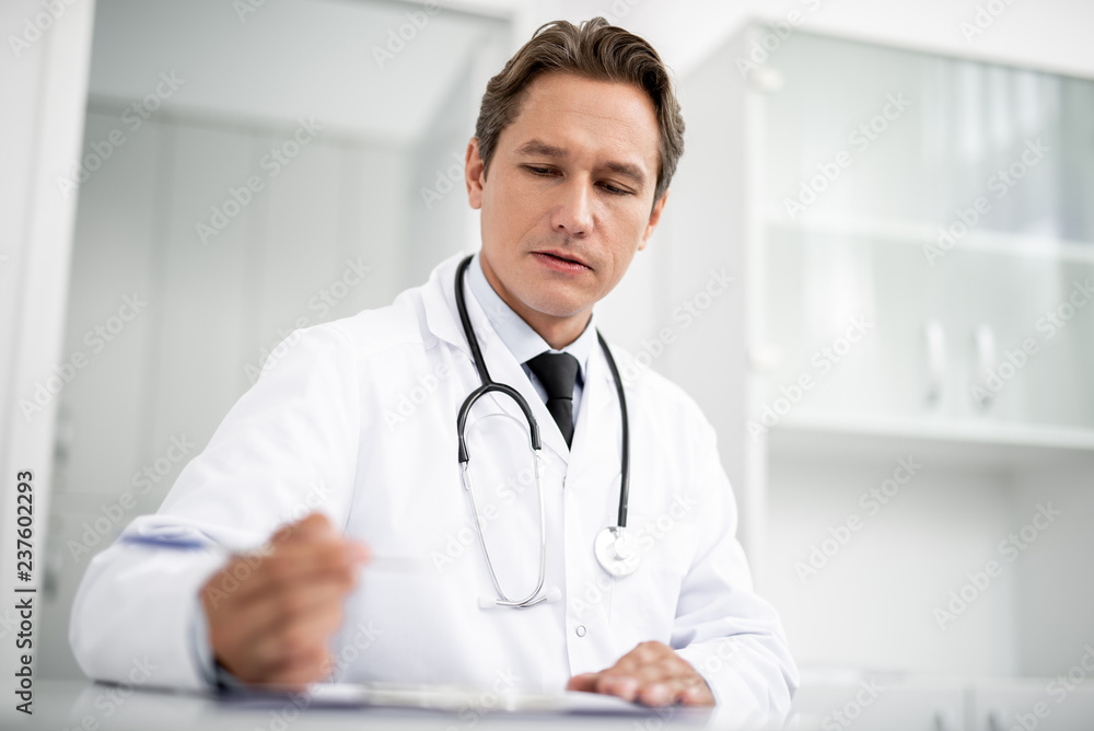 Thoughtful general practitioner looking attentively at the notes on his table and holding a pen