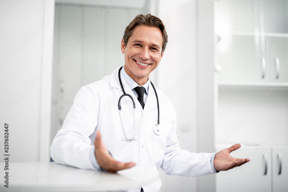 Cheerful enthusiastic general practitioner sitting with stethoscope on his neck and smiling happily while putting hands up Cheerful enthusiastic general practitioner sitting with stethoscope on his neck and smiling happily while putting hands up