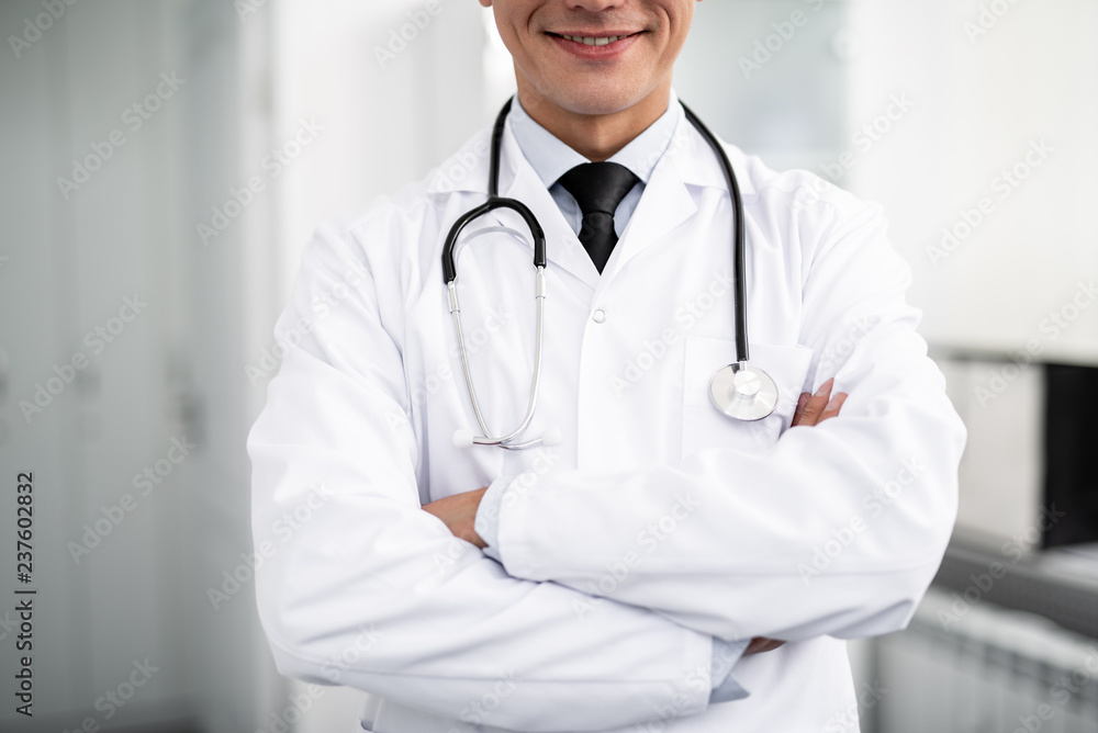 Cheerful enthusiastic doctor with stethoscope on his neck standing with his arms crossed and smiling happily