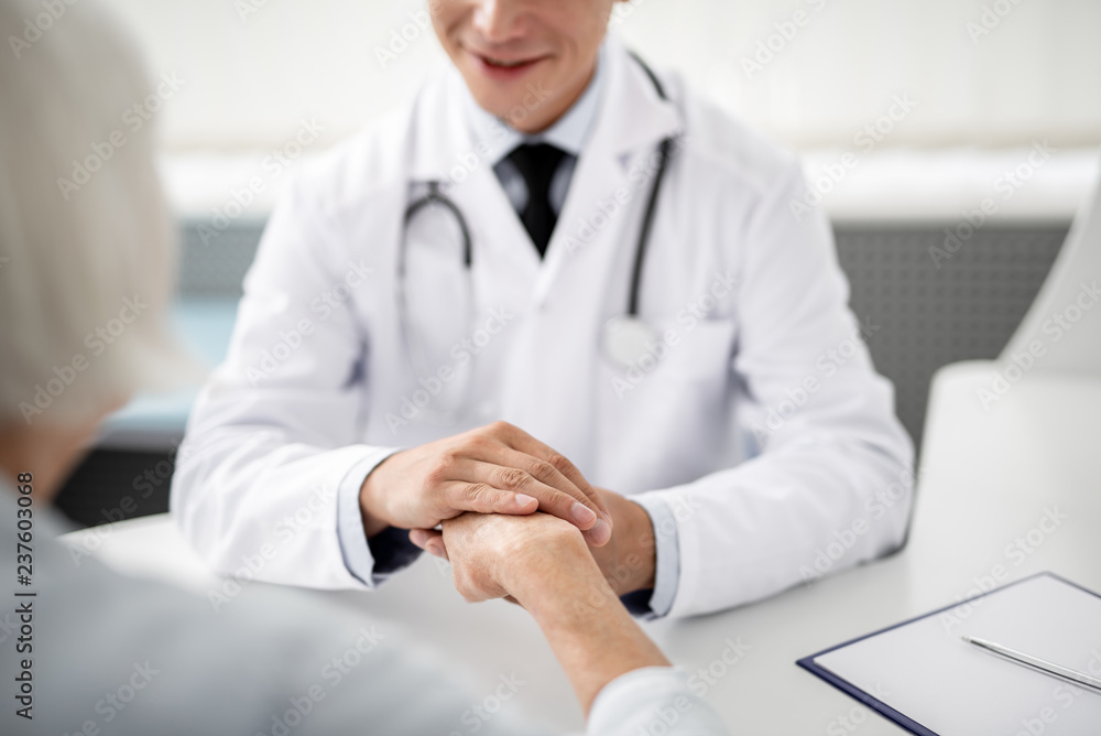 Kind smiling doctor sitting in front of his patient and gently touching her hand Kind smiling doctor sitting in front of his patient and gently touching her hand