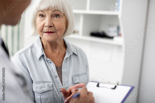 Calm elderly lady standing close to her doctor and looking attentively at him