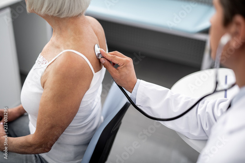 Careful experienced practitioner sitting next to his patient and using stethoscope while examining her