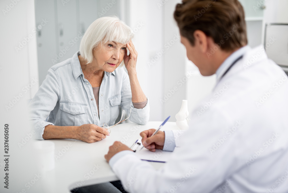 Serious elderly woman sitting in front of the general practitioner and looking at him while touching her head Serious elderly woman sitting in front of the general practitioner and looking at him while touching her head