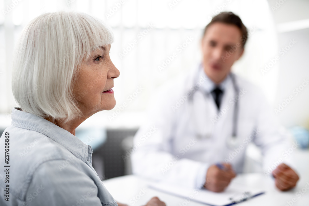 Close up of the face of aged woman thoughtfully looking into the distance with her practitioner sitting on the background Close up of the face of aged woman thoughtfully looking into the distance with her practitioner sitting on the background