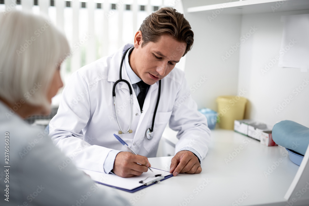 Attentive professional practitioner writing on the clipboard while sitting at the table with patient next to him Attentive professional practitioner writing on the clipboard while sitting at the table with patient next to him