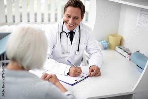 Positive emotional general practitioner sitting with the clipboard on the table and kindly smiling to his patient photo