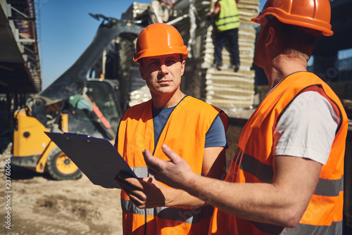 Calm attentive builder in bright uniform looking attentively at his coworker while standing outdoors with him and holding clipboard