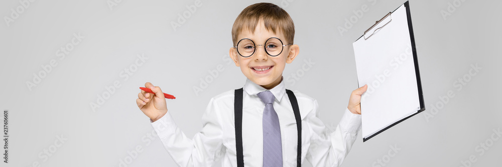 A charming boy in a white shirt, suspenders, a tie and light jeans stands on a gray background. The boy holds a pen and sheets for notes