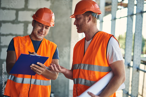 Enthusiastic builder holding clipboard and smiling while making notes and talking to colleague