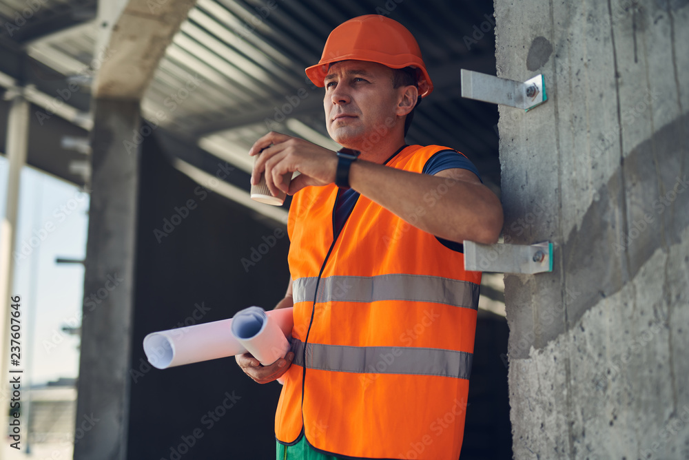 Calm experienced foreman in orange uniform thoughtfully drinking his coffee while being at work
