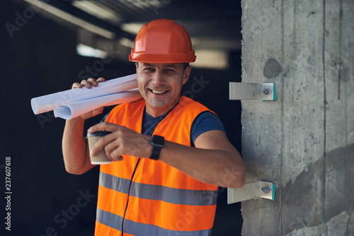 Positive emotional builder in bright orange uniform standing next to the wall and smiling while holding carton cup of coffee and drawings