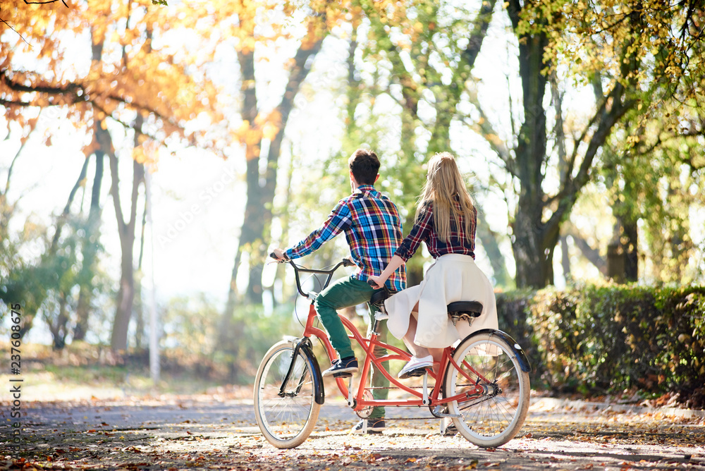 Back view of young tourist couple, man and blond long-haired woman cycling together tandem double red bicycle by park or forest sunny alley covered with golden leaves at autumn