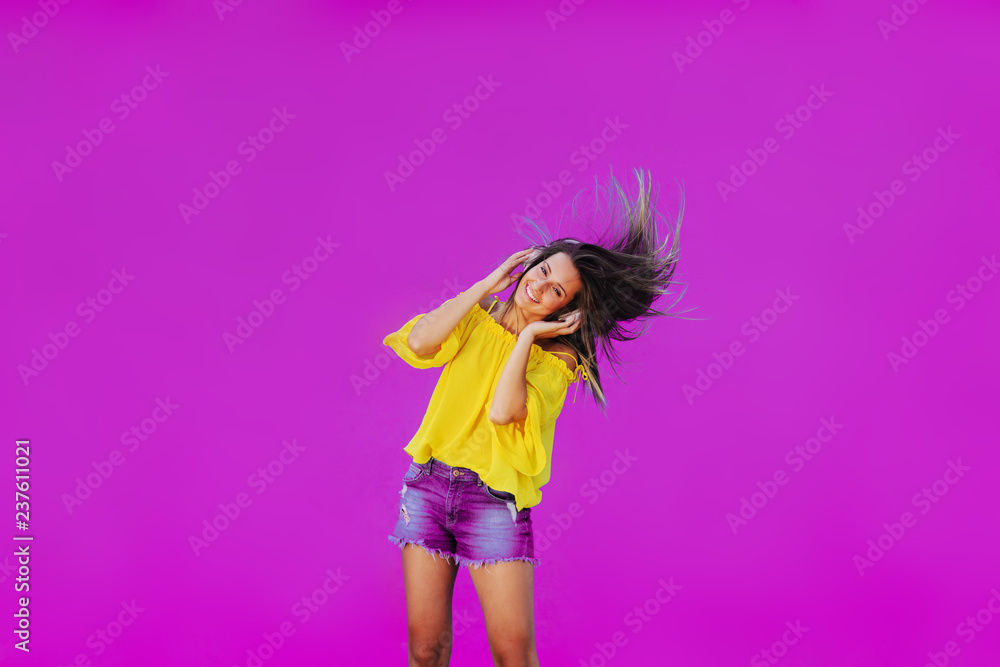 Cheerful girl in yellow shirt standing against pink wall with headset on her head. Dancing and smiling. Cheerful girl in yellow shirt standing against pink wall with headset on her head. Dancing and smiling.