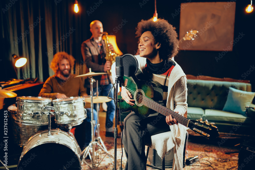 Fototapeta premium Mixed race woman singing and playing guitar while sitting on chair with legs crossed. In background drummer, saxophonist and bass guitarist.