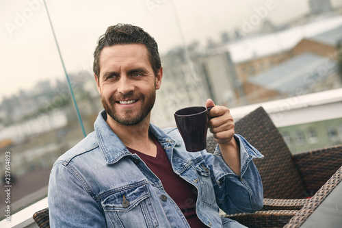 Waist up of adult smiling male with beard is spending time on cozy cafe and drinking coffee