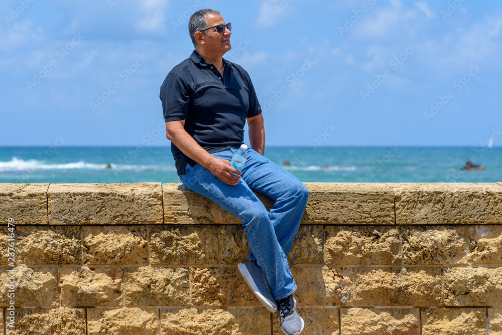 Smiling trendy tourist elderly man sitting by old brick historic stone.  Portrait of handsome aged fashion senior wearing black t-shirt and jeans on  blue sea and sky background. Vacation concept. Stock Photo