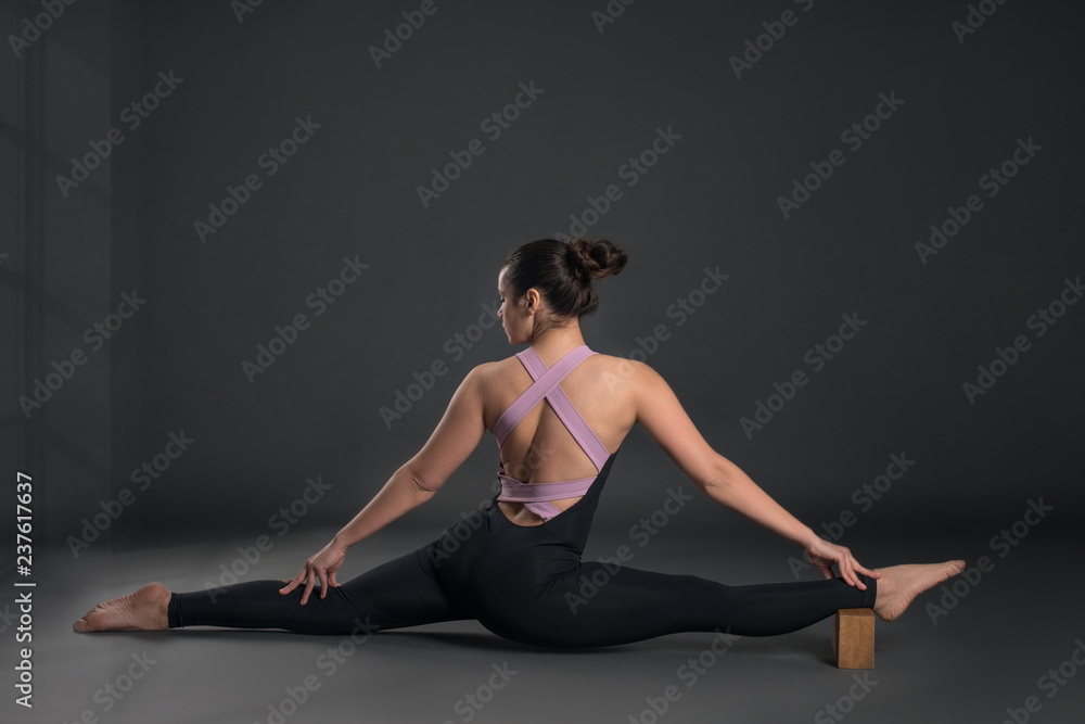 Young calm  woman  wearing black sportswear  does stretching exercise with brick, yoga exercise or pilates or stretching on gray background in studio, full length. 
