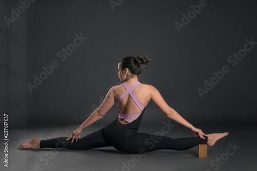 Young calm woman wearing black sportswear does stretching exercise with brick, yoga exercise or pilates or stretching on gray background in studio, full length. 