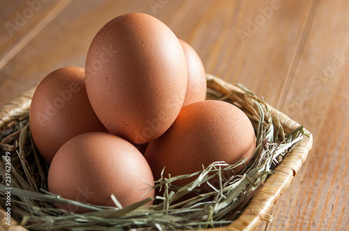 Five large brown eggs lie on a straw bedding in a wicker basket on a wooden table. Still life close-up.