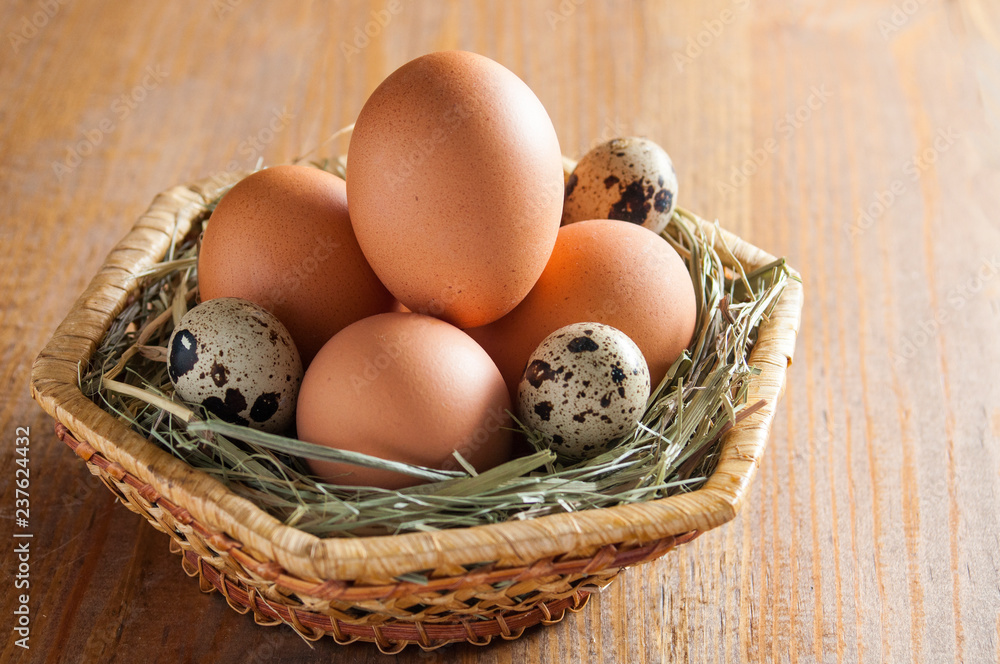 Chicken and quail eggs in a wicker basket, lying on the hay. On a wooden table. Chicken and quail eggs in a wicker basket, lying on the hay. On a wooden table.