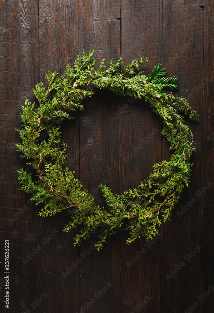 Christmas door wreath made of green fir branches on dark brown wooden background table or door with red and golden Christmas baubles and lights, copy space in center frame and empty mock up for text