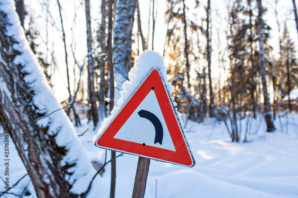 Red triangle road traffic sign indicating left move with flakes of snow, with the dawn sun on the background. Christmas frosty morning. Winter landscape, Tranquil nature in sunlight in park Red triangle road traffic sign indicating left move with flakes of snow, with the dawn sun on the background. Christmas frosty morning. Winter landscape, Tranquil nature in sunlight in park