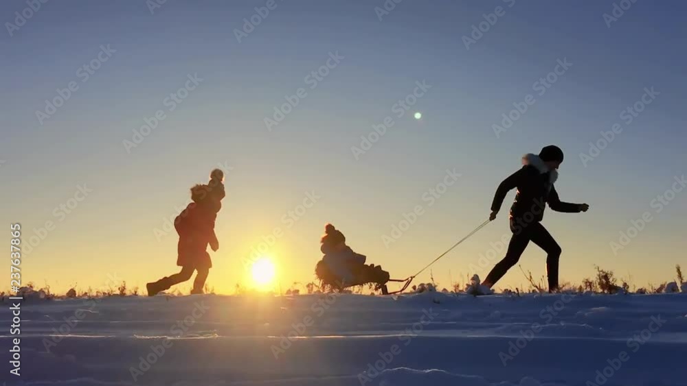 Teen daughter and mother pulling little son on sledge, running. Silhouette family playing in winter time. Active healthy lifestyle concept.