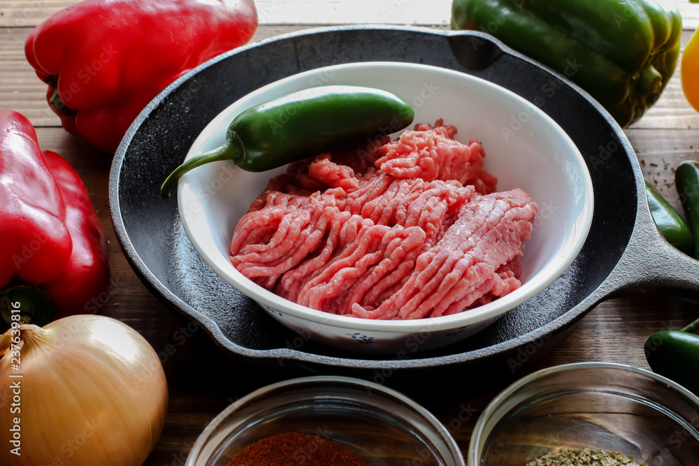 Raw ground beef inside skillet with peppers and other cooking ingredience around Raw ground beef inside skillet with peppers and other cooking ingredience around