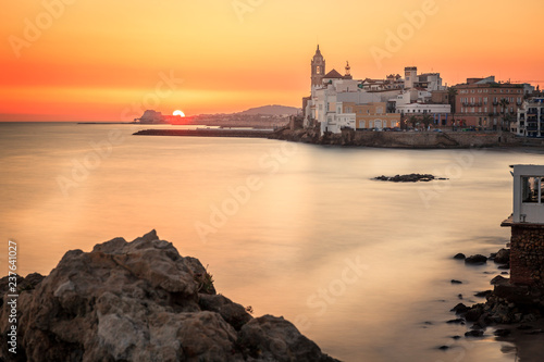 This is a sunset view with rocks on the foreground, the mediterranean sea blurried by a slow shutter speed and the town of sitges in the background.