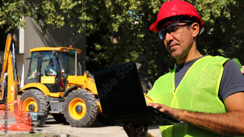Engineer using laptop in front of wheel loader excavator at construction site
