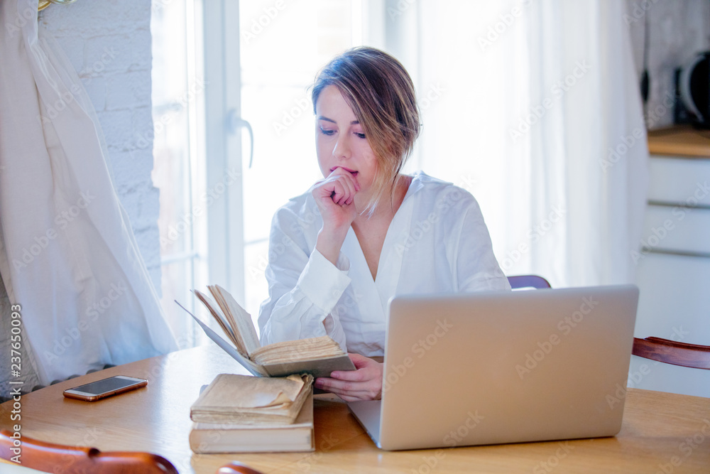 Young girl in white shirt sitting at table with books and laptop computer and looking for something. Business, education, shopping and other concept