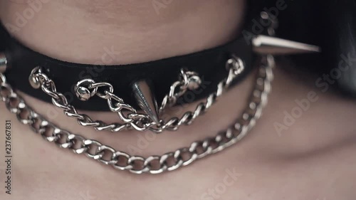Camera shows closeup of young brunette female's neck wearing thin black leather collar with silver chain plus hand with black nail polish on background of white walls.