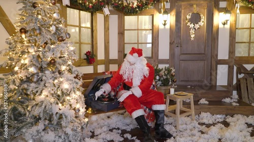 Santa Claus sits near the house between christmas trees, drinks milk, eats cookies, listens christmas songs on vinyl by gramophone, relaxes, enjoys, miss alone