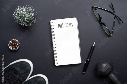 2019 new years resolutions sport flat lay composition. Black shoes, black dumbbells, glasses, blank notebook and Christmas decoration on black background. New year new healthy goals concept. 