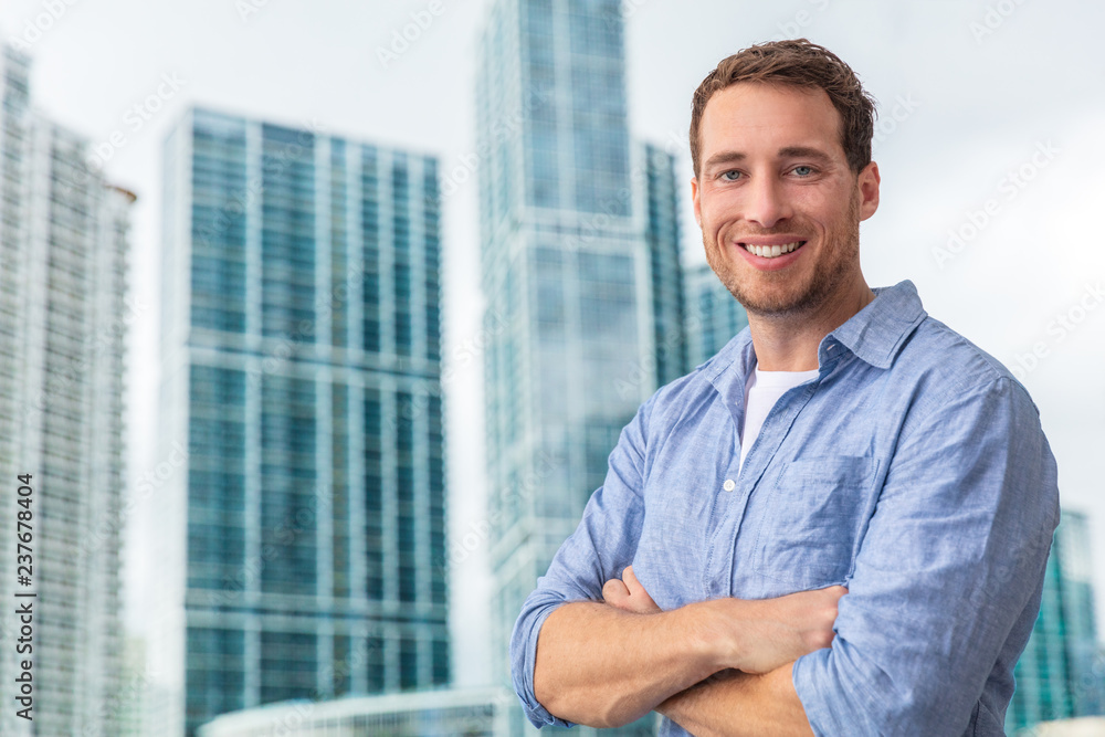 Happy young man businessman portrait young smiling confident in front of modern condo building skyscraper outside. City lifestyle people. Happy young man businessman portrait young smiling confident in front of modern condo building skyscraper outside. City lifestyle people.
